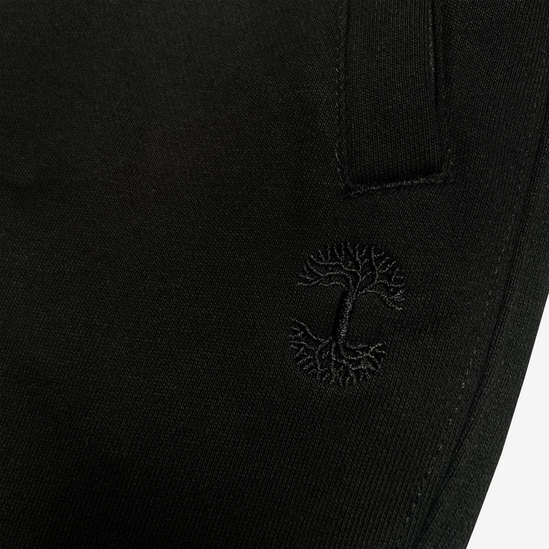Detailed close of embroidered Oaklandish tree logo under the front pocket of black joggers.