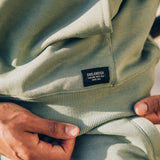 Detailed close-up Oaklandish label on the bottom left corner of the kangaroo pocket on an army green hoodie on a man sitting outside.