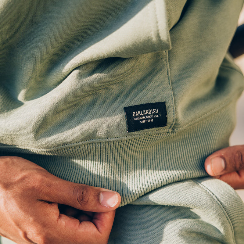 Detailed close up of woven label at bottom left corner of kangaroo pocket and ribbing at cuff and waistband of army standard pullover sweatshirt.