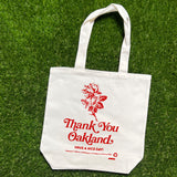 Cream shopping tote bag with red flower and the words “Thank-You Oakland. Have a Nice Day” lying on grass.