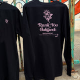 Long-sleeve black t-shirt with pink graphic of a flower with the words “Thank-You Oakland. Have a Nice Day” hanging outdoors on a fence.