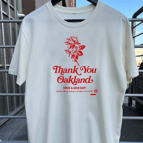 White t-shirt with a hand-drawn red flower and the words “Thank-You Oakland. Have a Nice Day” outdoors hanging on a fence.