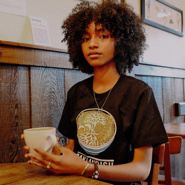 A woman in a cafe in a black t-shirt depicting a noodle bowl with an Oaklandish tree logo depicted in the noodles and a white Oaklandish wordmark. 