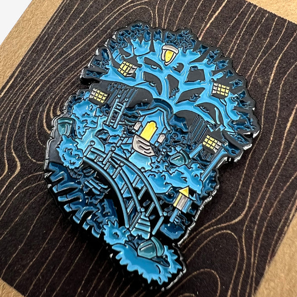 Close up of enamel pin with blue and yellow tree house Oaklandish tree logo on brown paper retail packaging. 