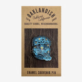 Enamel pin with blue and yellow tree house Oaklandish tree logo on brown paper retail packaging. 