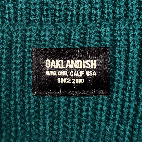 Detail close up of cuffed jade short knit beanie with Oaklandish square patch.