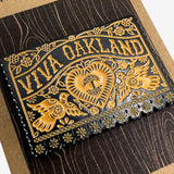 Close-up of gold and black enamel pin Viva Oakland wordmark with heart, birds and flowers on brown paper retail packaging. 