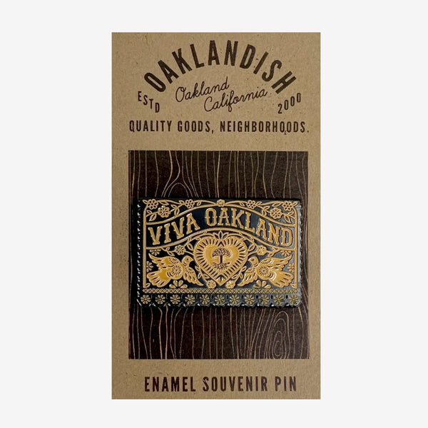 Gold and black enamel pin Viva Oakland wordmark with heart, birds and flowers on brown paper retail packaging. 