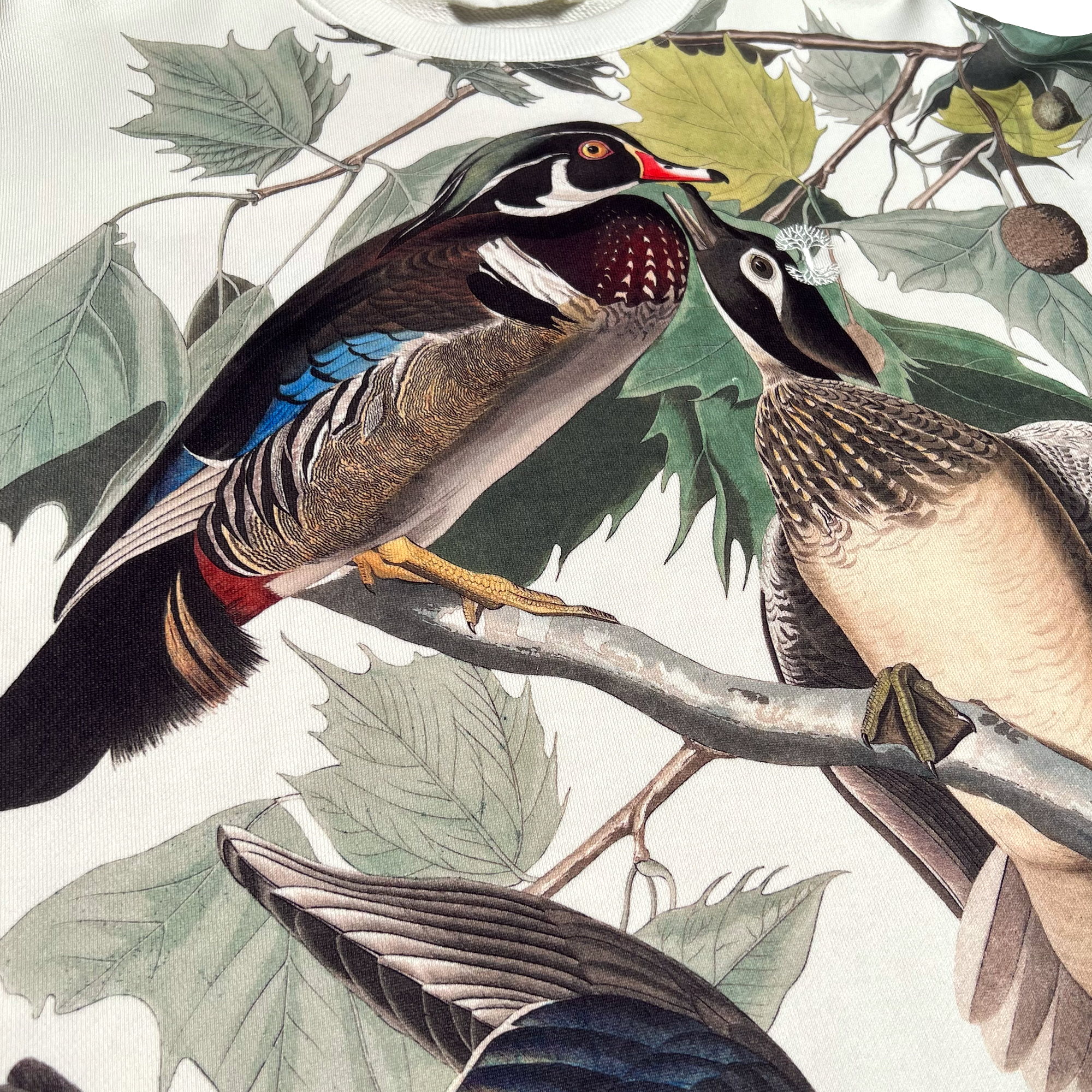 Detailed close up of wood duck all over print on cream crewneck sweatshirt with Oaklandish tree logo embroidered on left chest.