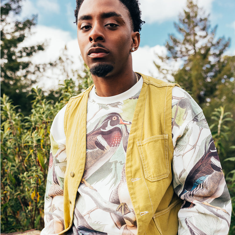 Man outdoors from the waist up wearing wood ducks all over print on cream crewneck Oaklandish sweatshirt. 