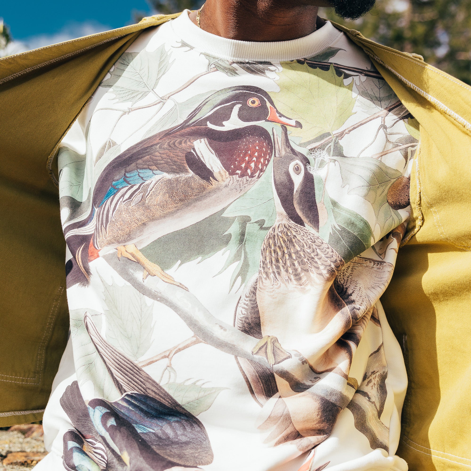 Close-up of a man’s chest wearing wood ducks all over print on cream crewneck Oaklandish sweatshirt. 