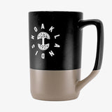 Side angle of the backside of a black and tan two-tone stoneware mug with the Oaklandish wordmark circling the Oaklandish tree logo.