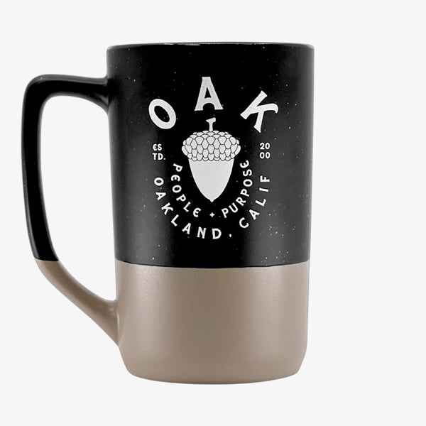 A black and tan two-tone stoneware mug depicting an acorn with the words “OAK People, Purpose, Oakland Calif.”