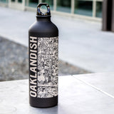 A black water bottle imprinted with an aerial Oakland map and Oakland wordmark, with a screw top on an outdoor ledge.