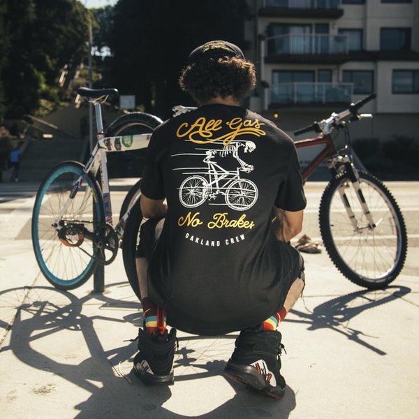 A man squatting on the sidewalk with bike wearing a black t-shirt with All Gas No Breaks, Oakland Crew design.