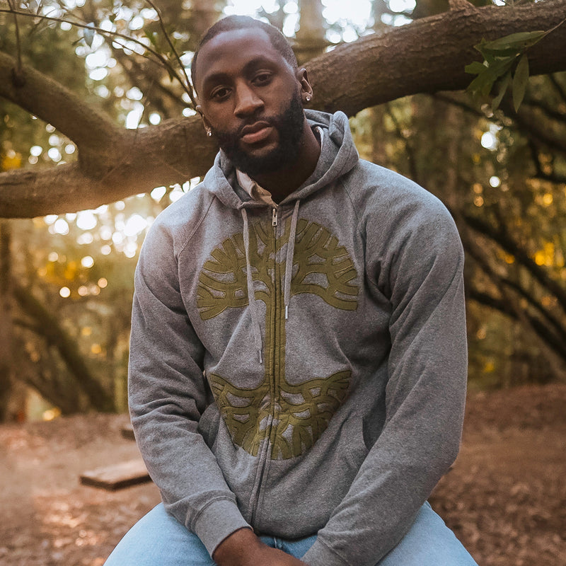 A man sitting in a park wearing a grey Oaklandish zip-up hoodie with an olive Oaklandish tree appliqué logo on the chest.