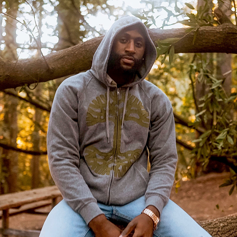 A man sitting in a park wearing a grey Oaklandish zip-up hoodie with hood up.
