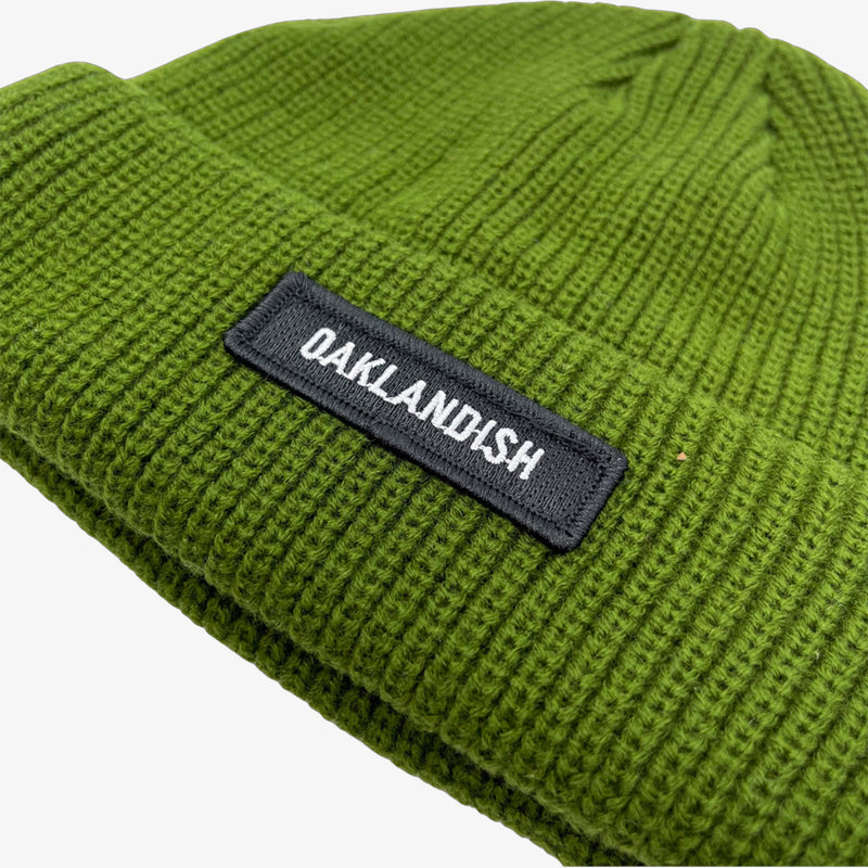 Close-up of shorter style olive green cuff beanie with white Oaklandish wordmark on a black patch on cuff.