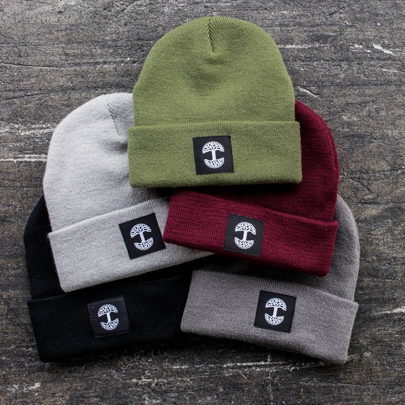 Five colors of Oaklandish logo beanie in olive green, dark grey, light grey, black, and burgundy on cement.