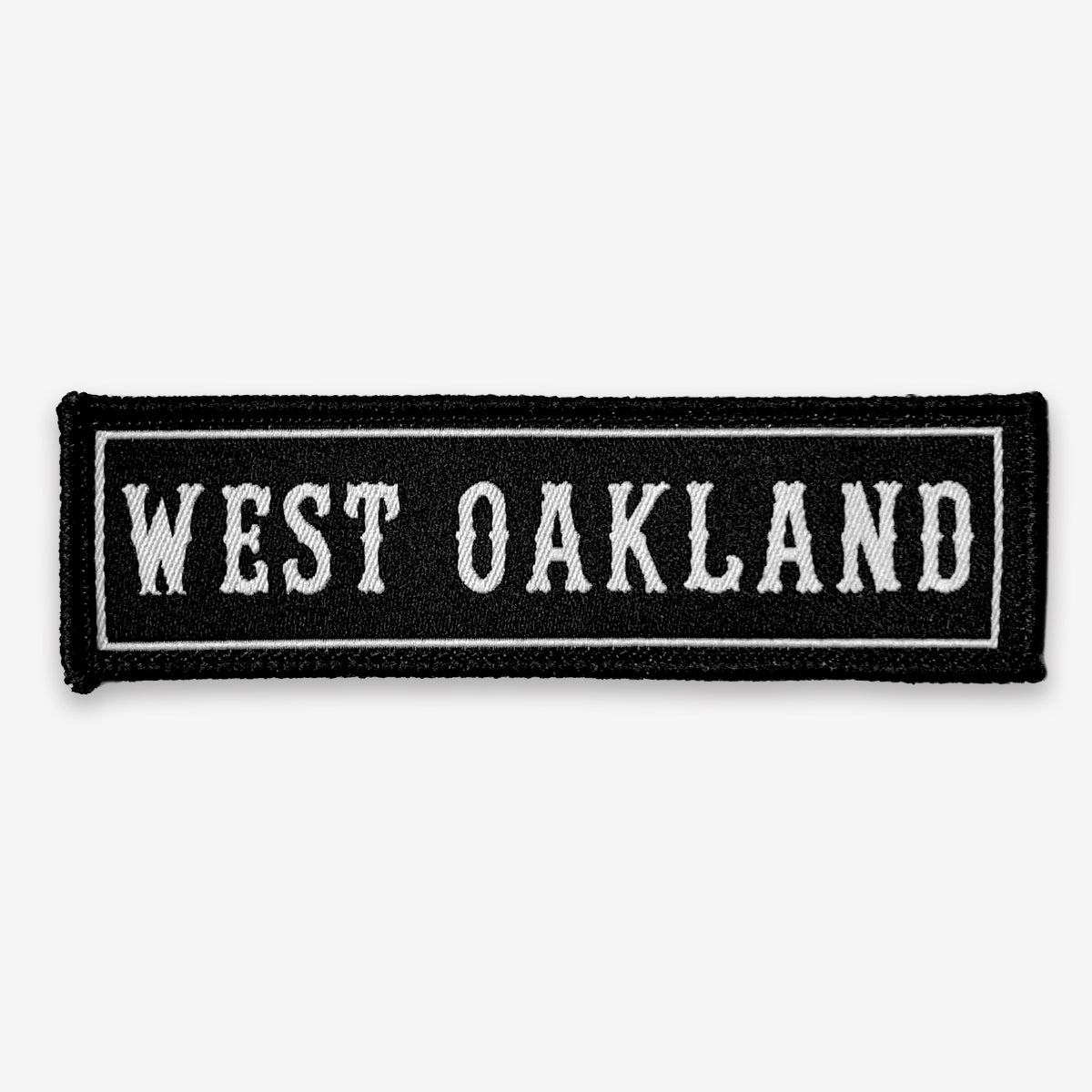 The West Oakland Patch by Oaklandish is a rectangular, black iron-on accessory with "WEST OAKLAND" embroidered in white, uppercase, bold letters. The centered text is framed by a thin white border, creating clean edges and a simple design.