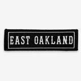 Black rectangle fabric iron patch, outlined with white line and capitalized word mark  EAST OAKLAND.”