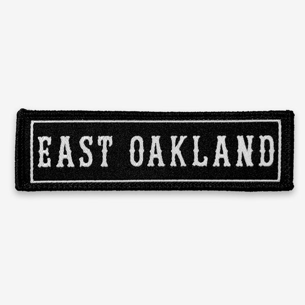 Black rectangle fabric iron patch, outlined with white line and capitalized word mark  EAST OAKLAND.”