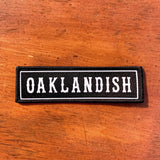 Rectangular black iron-on biker’s patch with white Oaklandish wordmark and white trim on wood background.