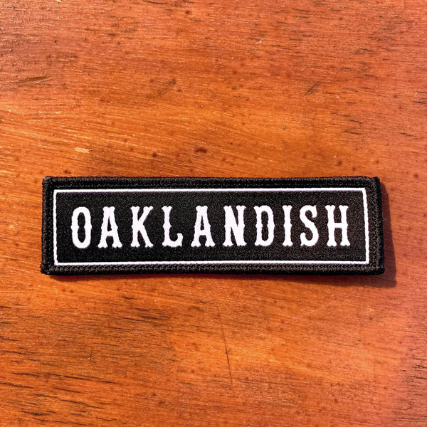 Rectangular black iron-on biker’s patch with white Oaklandish wordmark and white trim on wood background.