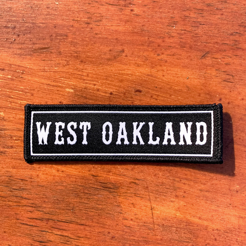 White WEST OAKLAND wordmark on a black rectangular biker-style patch on wood background.