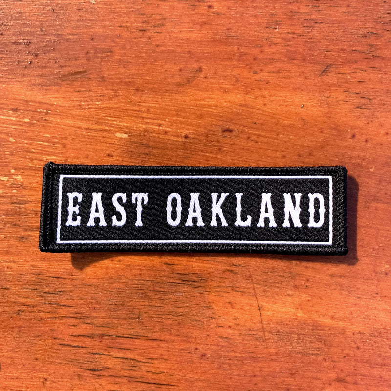 Black rectangle fabric iron patch, outlined with white line and capitalized word mark  EAST OAKLAND” on wood background.