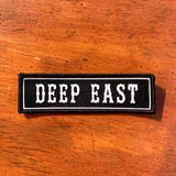 Black rectangle fabric iron patch, outlined with white line and capitalized word mark “DEEP EAST” on wood table.