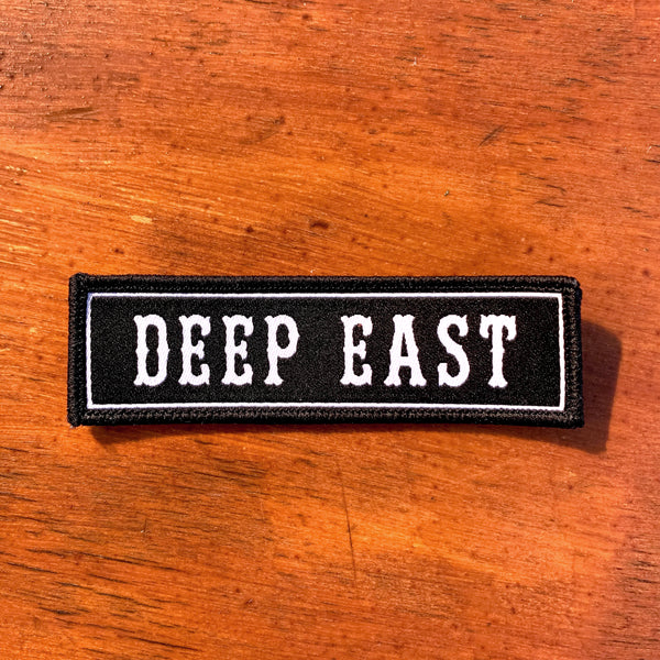 Black rectangle fabric iron patch, outlined with white line and capitalized word mark “DEEP EAST” on wood table.