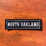 Black rectangle fabric iron patch, outlined with white line and capitalized word mark “NORTH OAKLAND" on wood table.