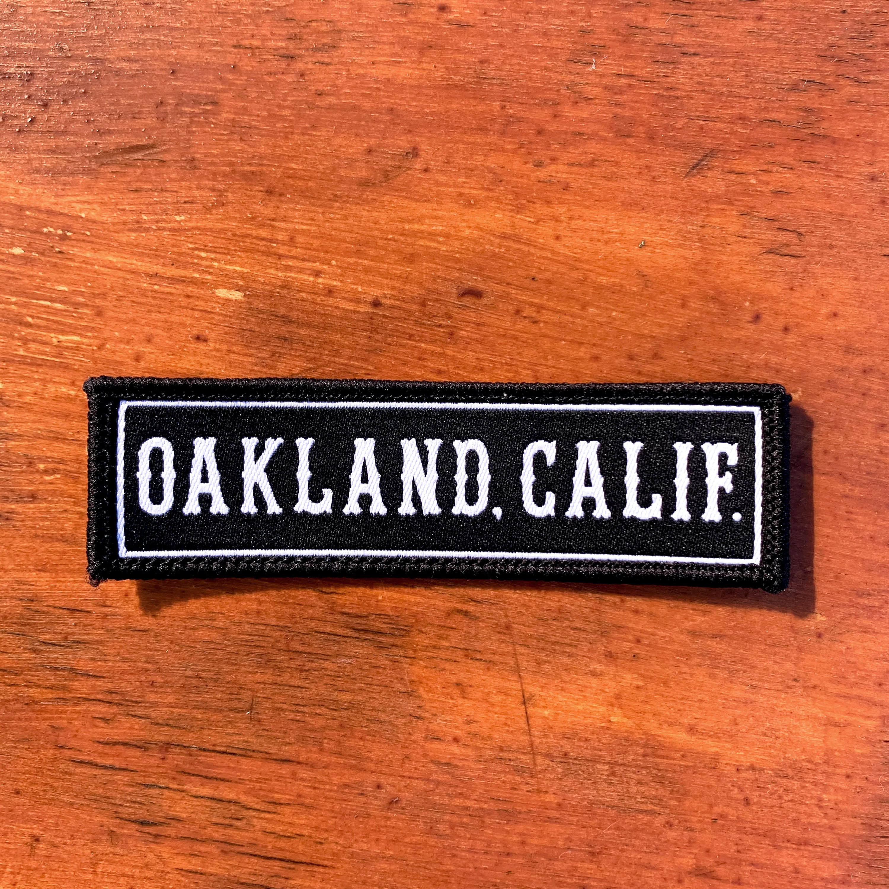 Black rectangle fabric iron patch, outlined with white line and capitalized word mark “OAKLAND. CALIF.” on wood desk.