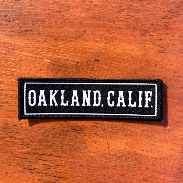 Black rectangle fabric iron patch, outlined with white line and capitalized word mark “OAKLAND. CALIF.” on wood desk.