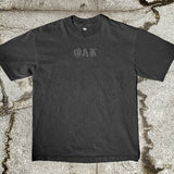 Vintage black t-shirt with black OAK wordmark in old style font with a bleed laying on asphalt.