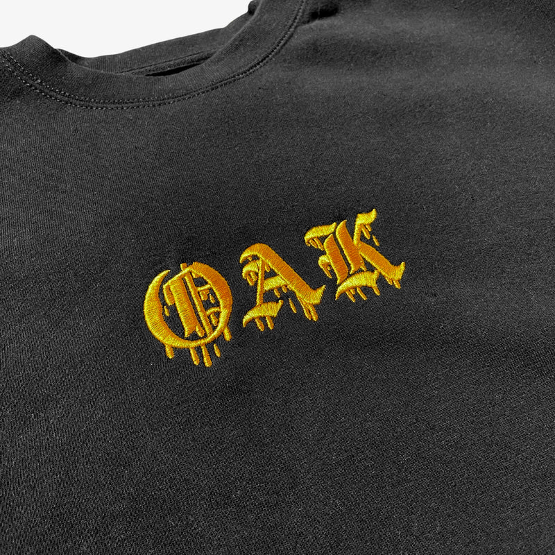Close-up of gold embroidered OAK in bleeding old style font on black crew neck sweatshirt.