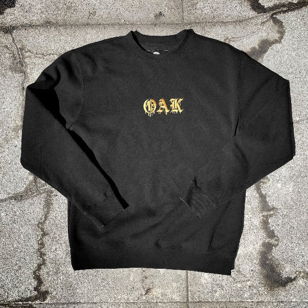 Black crew neck t-shirt with gold embroidered OAK in bleeding old style font lying on ashphalt.
