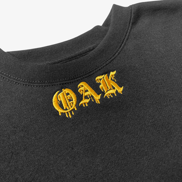 Close-up of gold embroidered OAK in bleeding old-style font on black crew neck t-shirt.
