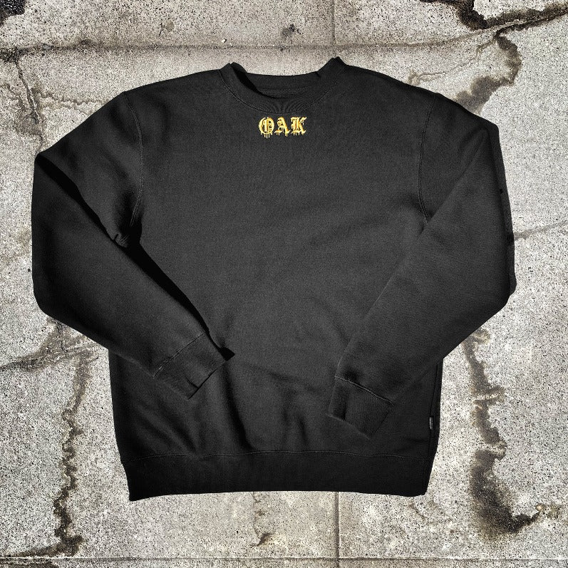 Black crew neck t-shirt with gold embroidered OAK in bleeding old-style font lying on the asphalt.