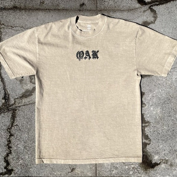 Mushroom brown t-shirt with black OAK wordmark in old style font with a bleed laying on asphalt.