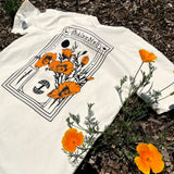 The back of a natural-color cotton t-shirt with California poppies, an Oaklandish wordmark, and tree logo lying on the ground.