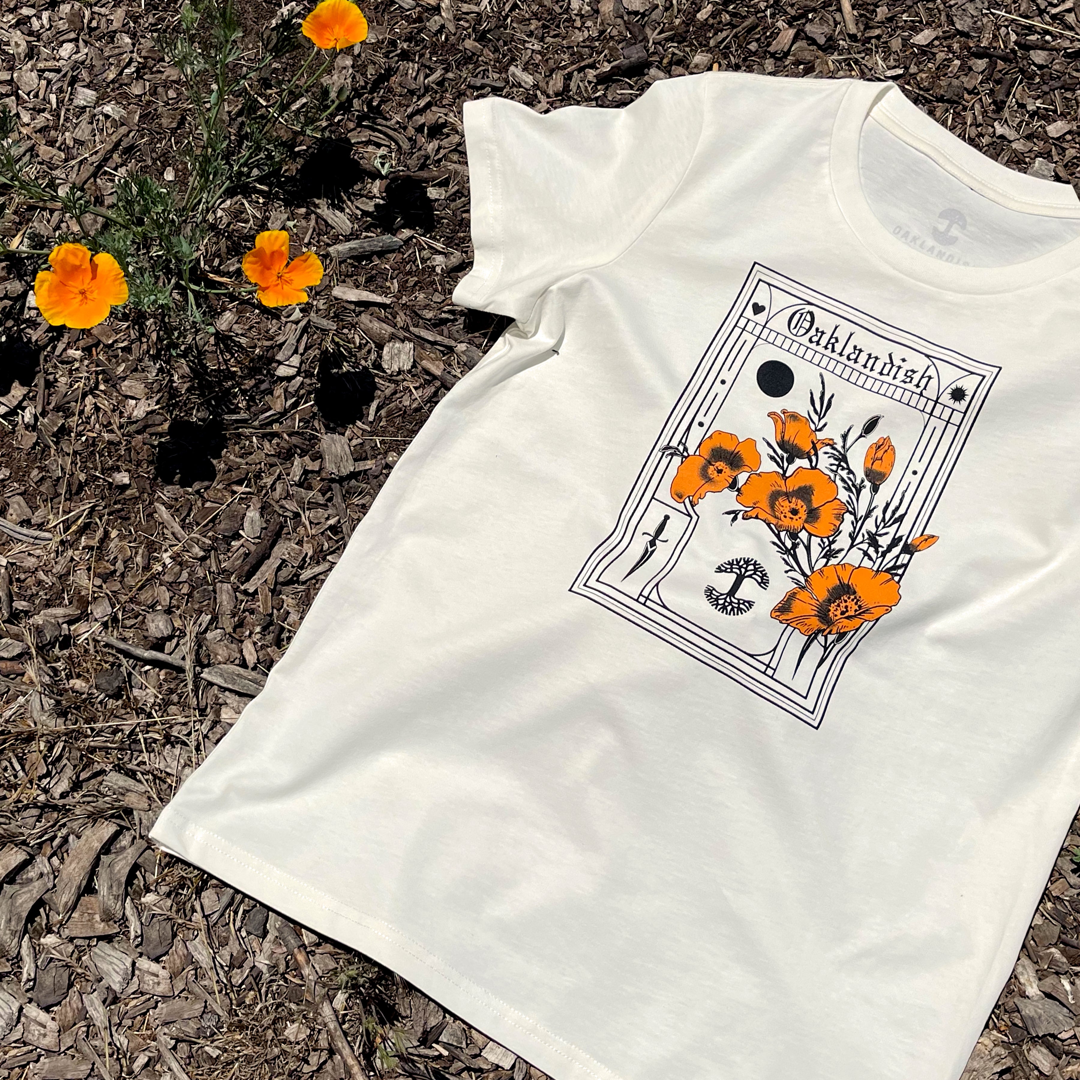 Women’s natural cotton t-shirt with California poppies, an Oaklandish wordmark, and tree logo. on the ground with real poppies.
