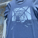 Close up the graphic of the eastern span of the Oakland bridge on the faded blue women’s t-shirt laying on metal.