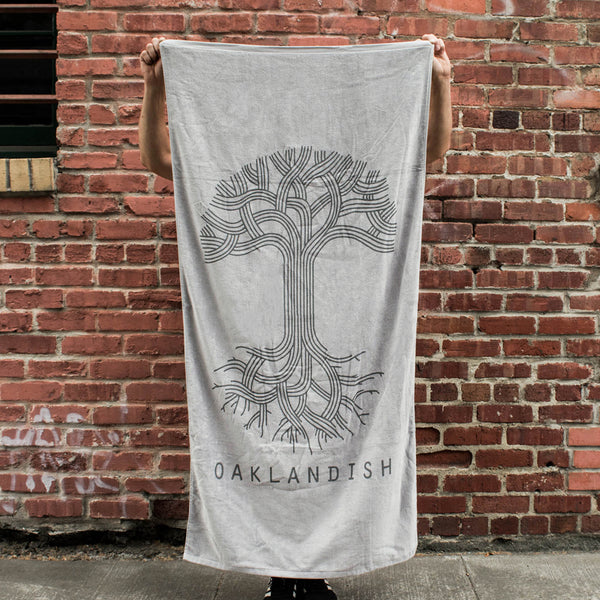 A person standing outside holding a plush oversized beach towel in silver grey with a classic Oaklandish logo and wordmark.