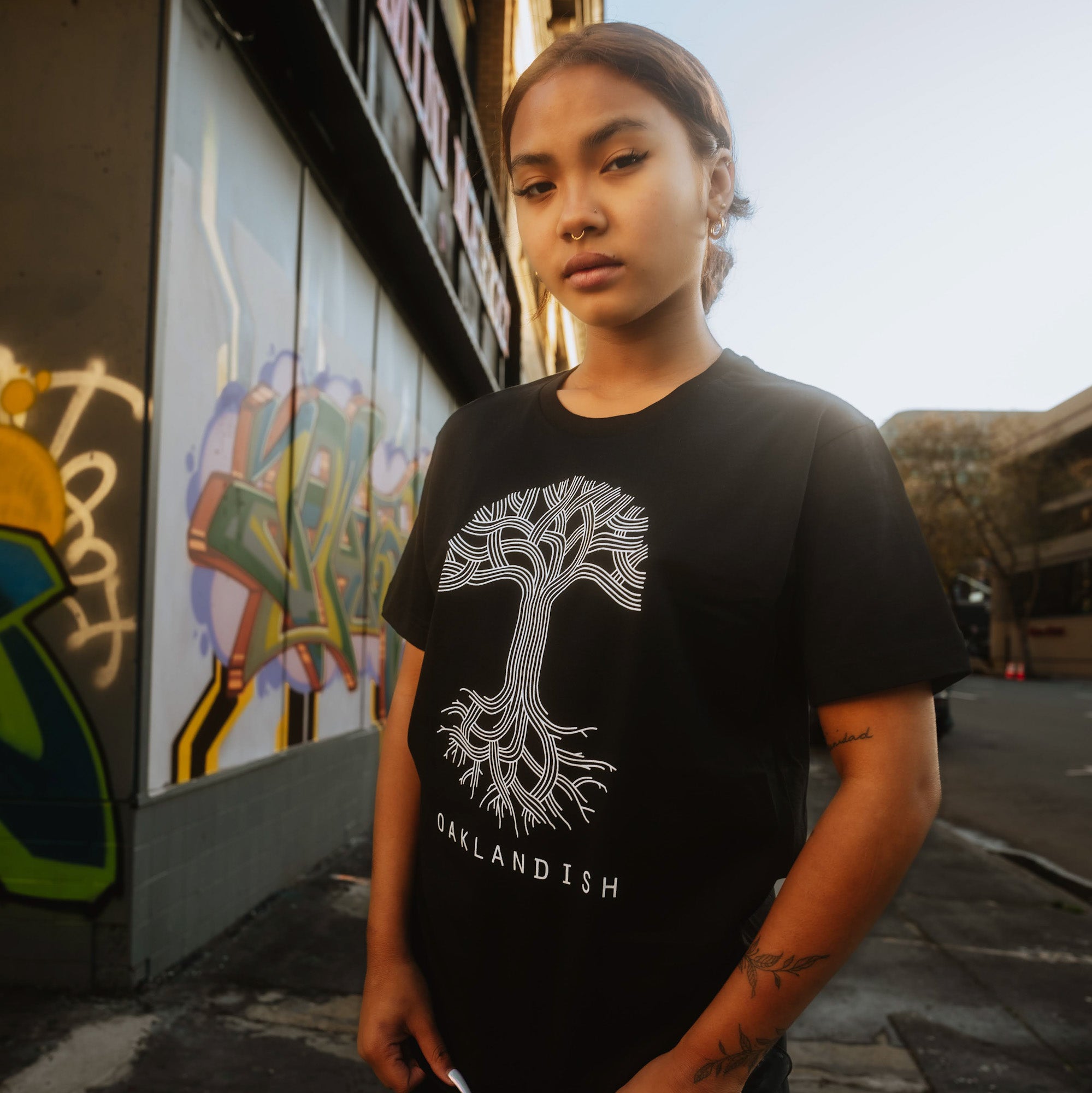 Female model wearing black t-shirt with a large white Oaklandish tree logo on the chest while standing outside.