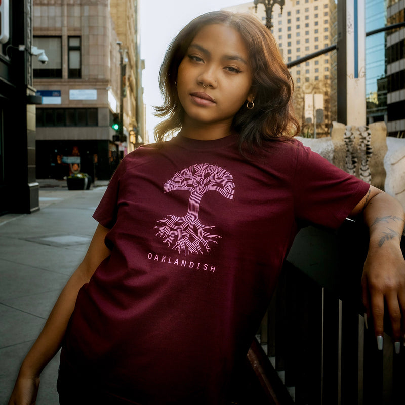 Female model wearing burgundy women’s cut tee with pink classic Oaklandish tree logo and wordmark on chest.