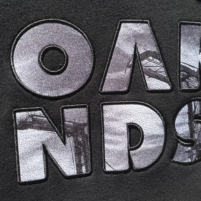 Close up of OAK capital letters with a photo of Oakland cranes in each letter on a black hooded sweatshirt.