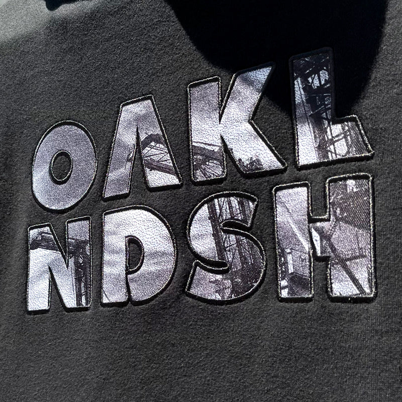 Close up of OAK capital letters with a photo of Oakland cranes in each letter on a black hooded sweatshirt.