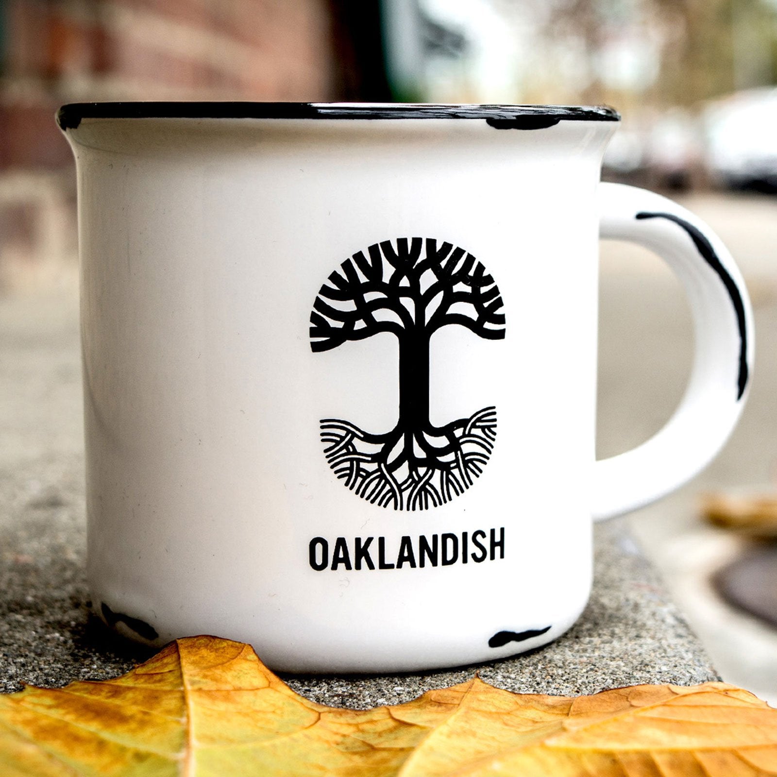 Front view of a white stoneware coffee mug featuring a retro style black Oaklandish tree logo on a cement table with a maple leaf.
